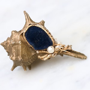 May include: A gold seashell with a blue velvet lining and a pearl accent. The seashell is a decorative object.
