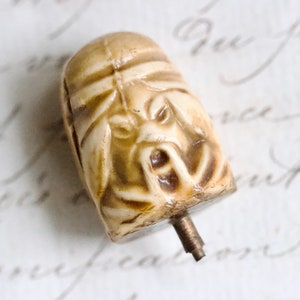 May include: A ceramic knob with a detailed face sculpted into the front. The knob has a metal post protruding from the bottom.