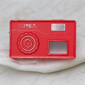 May include: A red plastic toy camera with the word "COMET" printed on the front. The camera has a viewfinder and a lens cover.