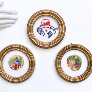 May include: Three gold-framed embroidery hoops with different designs. The first hoop features a woman with red hair wearing a red hat. The second hoop features a person in a blue robe with a yellow halo. The third hoop features a brown teddy bear wearing a red hat.