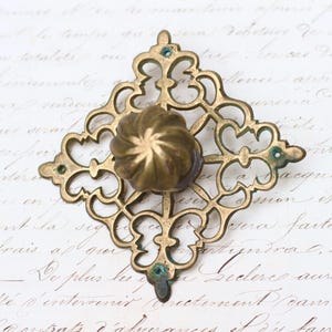 May include: A brass door knob with a decorative, ornate, square-shaped backplate. The backplate has a floral design with a raised center knob.