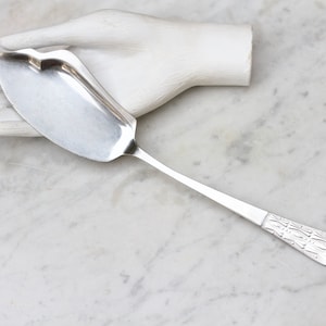 Puede incluir: Un utensilio de servir de plata con una hoja curva y un mango decorativo. El mango tiene un patrón repetitivo de líneas y formas.