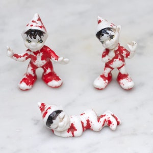 Peut inclure: Trois figurines en céramique d'elfes avec des éclaboussures de peinture rouge et blanche. Les elfes portent des chapeaux pointus et ont les cheveux noirs. Un elfe est debout les bras tendus, un autre est debout les bras croisés et un autre est couché.