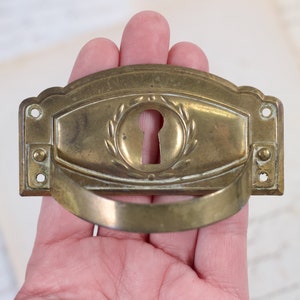 Op de afbeelding: Messing deurgreep met een sleutelgat en een decoratief kransontwerp. De greep heeft de vorm van een halve cirkel en een gladde, gepolijste afwerking.