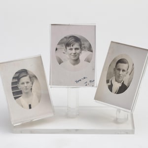 Pode incluir: Três fotografias preto e branco de homens jovens em molduras simples. As fotos são exibidas em um suporte de acrílico transparente. A foto à esquerda tem a inscrição "Yours, J.R."