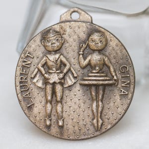 Puede incluir: Un amuleto de metal de color bronce con dos figuras, un niño y una niña, en relieve. El niño lleva un disfraz y la niña lleva un tutú. El amuleto está inscrito con los nombres "LAURENT" y "GINA".