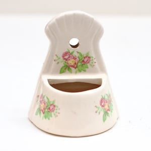 May include: White ceramic wall mounted toothbrush holder with a floral design featuring pink roses and green leaves.