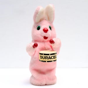 May include: A vintage pink plush bunny toy with white inner ears, green eyes, and red nose and accents. The bunny holds a small drum with the word "DURACEL" printed on it. The toy is standing upright.