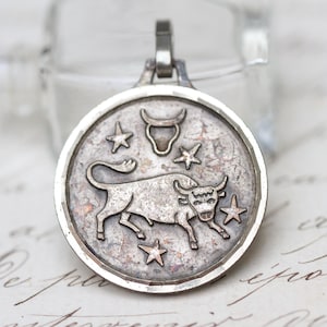 May include: A silver pendant depicting the Taurus zodiac sign. The circular pendant features a raised image of a bull, stars, and the Taurus symbol. It hangs from a small loop at the top.