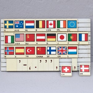 Pode incluir: Um quadro branco com uma grade de quadrados pequenos, cada um contendo uma bandeira que representa um país diferente. As bandeiras estão organizadas em ordem alfabética pelo nome do país. As bandeiras incluem Suécia, Austrália, Países Baixos, Bélgica, Canadá, Irlanda, União Europeia, Itália, Estados Unidos, União Soviética, Áustria, Alemanha, Japão, Portugal, França, Noruega, Espanha, Turquia, China, Finlândia, Reino Unido, Luxemburgo, Suíça e Dinamarca.