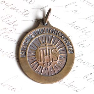 Puede incluir: Una medalla religiosa de bronce con un dise&ntilde;o circular. La medalla presenta un dise&ntilde;o de rayos de sol con las letras "IHS" en el centro. La inscripci&oacute;n alrededor del borde dice "MARIA CONGREGATIO".
