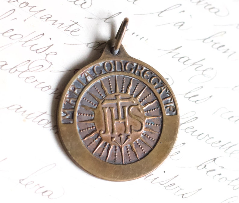 Puede incluir: Una medalla religiosa de bronce con un dise&ntilde;o circular. La medalla presenta un dise&ntilde;o de rayos de sol con las letras "IHS" en el centro. La inscripci&oacute;n alrededor del borde dice "MARIA CONGREGATIO".