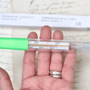 Puede incluir: Un termómetro de vidrio transparente y verde con una escala grande en grados Celsius. El termómetro tiene una carcasa de plástico verde y un tubo de vidrio transparente con un líquido rojo en su interior. El termómetro está etiquetado con "Termometro Escala Larga Escala Celsius C" y "CE".