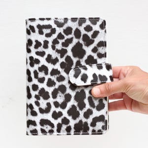 May include: A black and white leopard print planner with a snap closure. The planner is held by a hand against a white background. The cover features a repeating pattern of black spots on a white background.