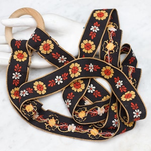 May include: Black ribbon with a floral pattern in red, yellow, and white, with a wooden ring. The ribbon has a gold trim and is arranged in a decorative pattern. The floral design is repeated along the length of the ribbon.