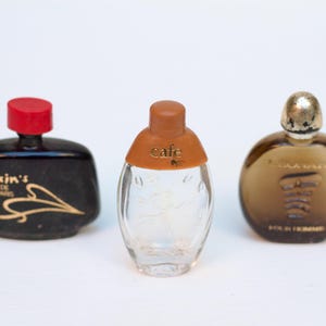Pode incluir: Três frascos de perfume em miniatura. O frasco à esquerda é preto com letras douradas que dizem "Maxim's" e uma tampa vermelha. O frasco do meio é de vidro transparente com uma tampa marrom e letras douradas que dizem "Cafe". O frasco à direita é de vidro marrom com uma tampa dourada.