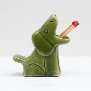 Könnte beinhalten: Ein grüner Keramik-Alligator-Streichholzhalter mit zwei Streichhölzern im Maul. Der Alligator hat eine hellgrüne Farbe mit glänzendem Finish. Die Streichhölzer haben rote Spitzen.