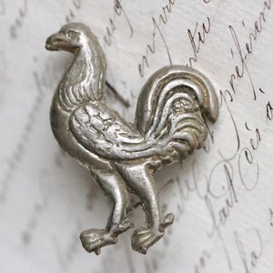 Puede incluir: Un broche de gallo plateado con detalles elaborados. El gallo está de pie con las alas extendidas y las plumas de la cola desplegadas.