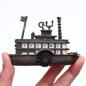 Op de afbeelding: Een klein bruin en crèmekleurig stoombootmodel. Het heeft een schoepenrad, een schoorsteen en een vlag. De woorden "Japan Made" zijn zichtbaar. Het model wordt in een hand gehouden. Miniatuur replica.