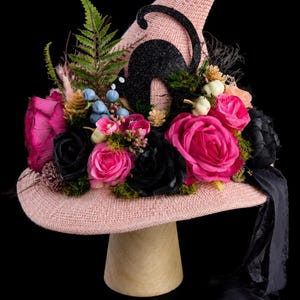 May include: A light pink knit pointed hat adorned with a black glittery cat silhouette, vibrant fuchsia and black artificial roses, green fern fronds, small blue berries, tiny white pumpkins, moss accents, and a long black fabric ribbon.