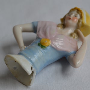 Pode incluir: Uma figurinha de porcelana de uma mulher vestindo um vestido azul e rosa com um detalhe de flor amarela. Ela est&aacute; usando um chap&eacute;u amarelo com uma borda branca. A figurinha est&aacute; em uma pose reclinada.