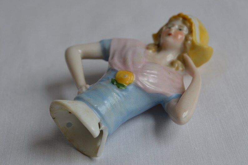Pode incluir: Uma figurinha de porcelana de uma mulher vestindo um vestido azul e rosa com um detalhe de flor amarela. Ela est&aacute; usando um chap&eacute;u amarelo com uma borda branca. A figurinha est&aacute; em uma pose reclinada.