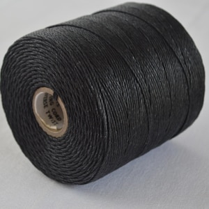 May include: A spool of black cord, tightly wound and cylindrical in shape. The cord appears to be made of a woven material. The spool has a cardboard core with printed text. The background is a plain, light surface.