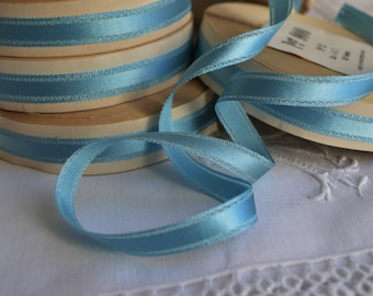 May include: A spool of light blue satin ribbon with a white edge. The ribbon is wrapped around a wooden spool and is on a white surface.