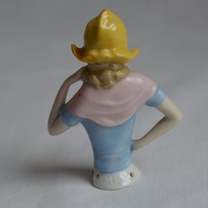 Pode incluir: Uma boneca de porcelana para alfinetes com um chap&eacute;u amarelo, um len&ccedil;o rosa e um vestido azul. A boneca est&aacute; de p&eacute; de costas para a c&acirc;mera e tem um buraco nas costas para os alfinetes.