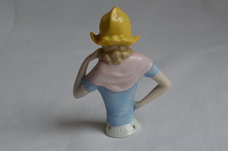 Pode incluir: Uma boneca de porcelana para alfinetes com um chap&eacute;u amarelo, um len&ccedil;o rosa e um vestido azul. A boneca est&aacute; de p&eacute; de costas para a c&acirc;mera e tem um buraco nas costas para os alfinetes.