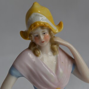 Pode incluir: Uma figura de porcelana de uma mulher usando um chap&eacute;u amarelo e branco e um vestido rosa e azul. A figura est&aacute; sentada com um bra&ccedil;o levantado.