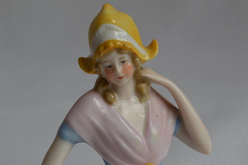 Pode incluir: Uma figura de porcelana de uma mulher usando um chap&eacute;u amarelo e branco e um vestido rosa e azul. A figura est&aacute; sentada com um bra&ccedil;o levantado.