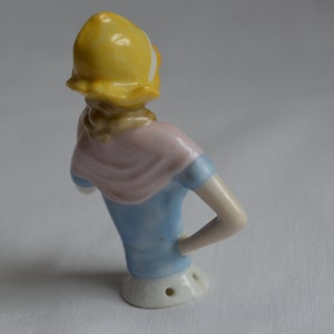 Pode incluir: Uma figurinha de porcelana de uma mulher usando um chap&eacute;u amarelo, um len&ccedil;o rosa e um vestido azul. A figurinha est&aacute; em p&eacute; com um bra&ccedil;o levantado e o outro bra&ccedil;o dobrado no cotovelo.