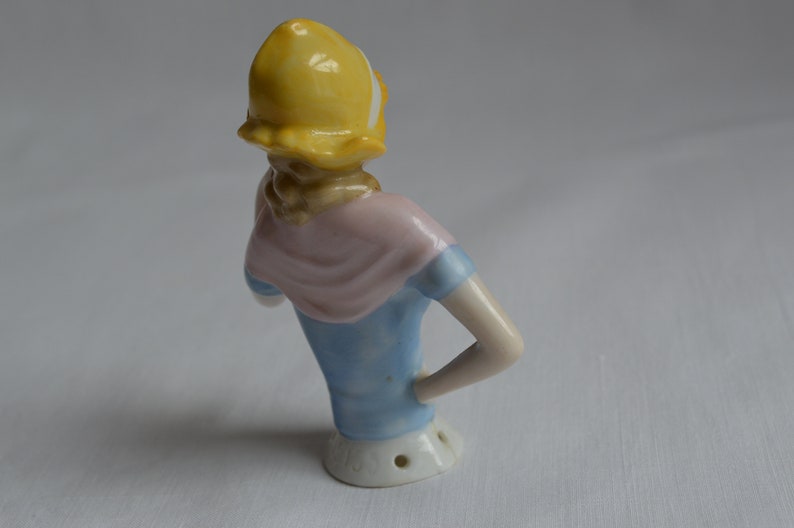 Pode incluir: Uma figurinha de porcelana de uma mulher usando um chap&eacute;u amarelo, um len&ccedil;o rosa e um vestido azul. A figurinha est&aacute; em p&eacute; com um bra&ccedil;o levantado e o outro bra&ccedil;o dobrado no cotovelo.