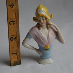Pode incluir: Uma boneca de porcelana para alfinetes com um chap&eacute;u amarelo, um xale rosa e um vestido azul. A boneca tem cabelo loiro e est&aacute; segurando a m&atilde;o no queixo.