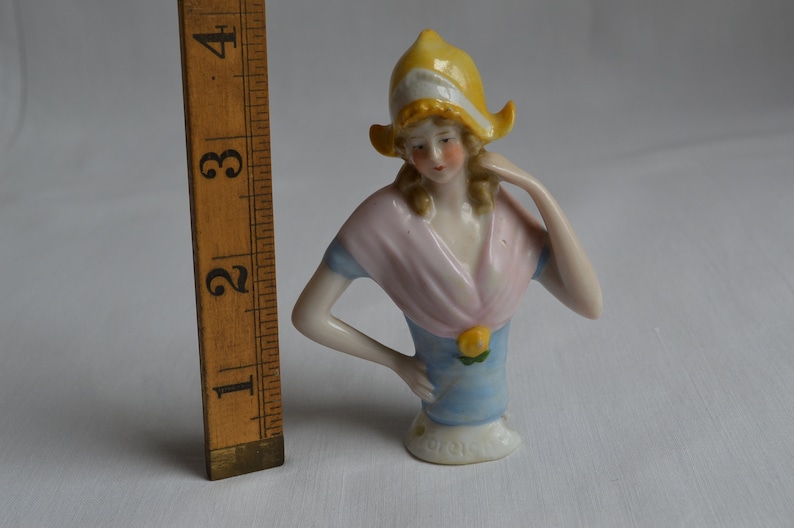 Pode incluir: Uma boneca de porcelana para alfinetes com um chap&eacute;u amarelo, um xale rosa e um vestido azul. A boneca tem cabelo loiro e est&aacute; segurando a m&atilde;o no queixo.