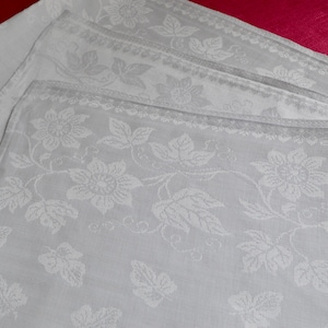 May include: A set of white linen napkins with a delicate floral pattern. The napkins are folded and stacked on top of each other.