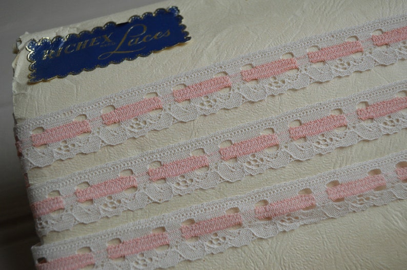 May include: Three rows of white lace trim with pink stripes. The trim is on a white background with a blue label that says "Wentex Laces".