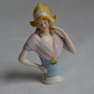 Pode incluir: Uma boneca de alfinete de porcelana com um chap&eacute;u amarelo, um xale rosa e um vestido azul. A boneca tem cabelo loiro e est&aacute; segurando a m&atilde;o no queixo. A base do alfinete &eacute; marcada com "Dollcraft".