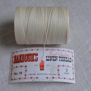 May include: A cream-coloured spool of linen thread is positioned above a label. The label reads "BARBOUR'S BEST QUALITY LINEN THREAD", with "No. 18" and "3 CORD" details. The label also states the thread is made in Lisburn, Northern Ireland.