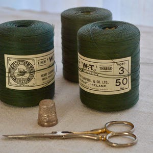 Op de afbeelding: Drie spoelen groene linnen draad met witte etiketten. De etiketten hebben de tekst "W.T. THREAD" en "HENRY CAMPBELL & CO. LTD. BELFAST, IRELAND".
