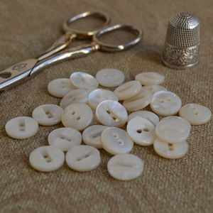 May include: A pile of white pearl buttons with two holes each, scattered on a beige fabric surface. A pair of silver scissors and a silver thimble are also visible.