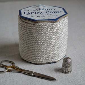 May include: A spool of white lacing cord with a blue label that reads "Extra Quality Lacing Cord Col. White No. 101X 36 yds. Made in England". There are also a pair of silver scissors and a silver thimble.