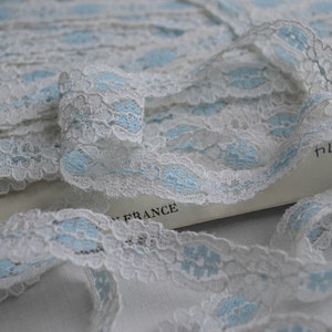 May include: White and light blue lace trim with a delicate floral pattern. The lace is folded and arranged on a white surface.