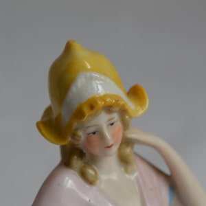 Pode incluir: Uma figura de porcelana de uma mulher usando um chap&eacute;u amarelo e branco. Ela est&aacute; usando um vestido rosa e tem cabelo loiro.