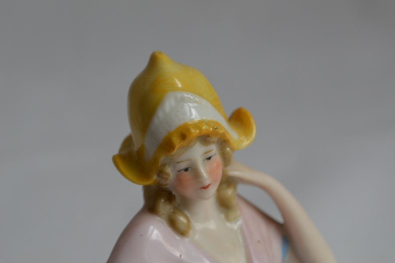 Pode incluir: Uma figura de porcelana de uma mulher usando um chap&eacute;u amarelo e branco. Ela est&aacute; usando um vestido rosa e tem cabelo loiro.