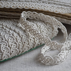 May include: White braided trim with a decorative looped pattern. The trim is folded and stacked on a white surface.