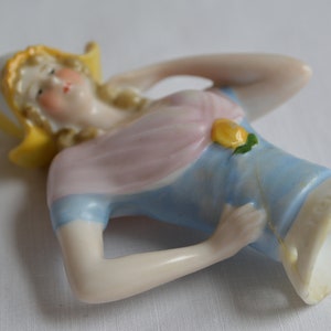Pode incluir: Um busto de boneca de porcelana com um chap&eacute;u amarelo, um vestido rosa e azul e um detalhe de flor amarela. A boneca est&aacute; em uma pose reclinada com um bra&ccedil;o levantado em dire&ccedil;&atilde;o &agrave; cabe&ccedil;a.