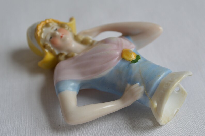 Pode incluir: Um busto de boneca de porcelana com um chap&eacute;u amarelo, um vestido rosa e azul e um detalhe de flor amarela. A boneca est&aacute; em uma pose reclinada com um bra&ccedil;o levantado em dire&ccedil;&atilde;o &agrave; cabe&ccedil;a.