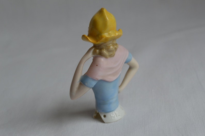 Pode incluir: Uma boneca de porcelana com um chap&eacute;u amarelo, um len&ccedil;o rosa e um vestido azul. A boneca est&aacute; faltando a parte inferior do corpo e tem uma marca na parte inferior que diz "1514".